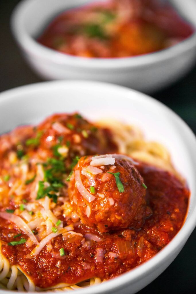 A close-up of a bowl of spaghetti topped with tomato sauce, meatballs, grated cheese, and chopped herbs. Another similar bowl is blurred in the background.