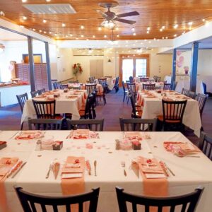 A banquet hall decorated for a celebration, with tables covered in white cloths and pink accents, set with plates, glasses, and utensils. String lights hang from the wooden ceiling, creating a warm, festive atmosphere.