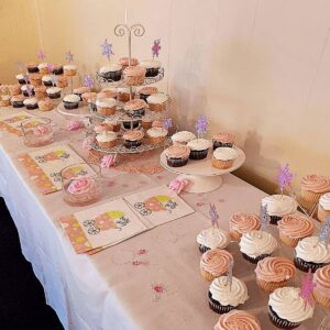 A dessert table decorated for a baby shower, featuring pink and white frosted cupcakes, some with glittery star toppers, napkins with baby carriage designs, and glass bowls with pink decorations on a white tablecloth.