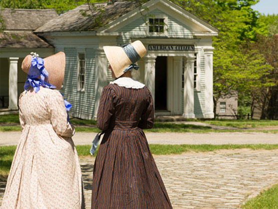 Two women in 19th-century dresses and bonnets stand on a cobblestone path, facing a white historic building with a sign that reads American Citizen. Trees and grass surround the scene.