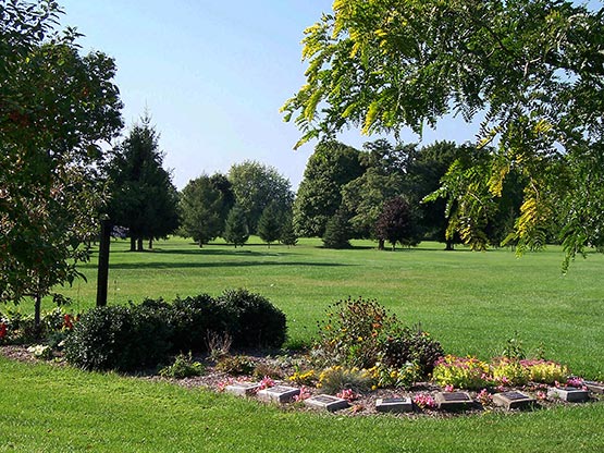 A peaceful cemetery scene with flat grave markers surrounded by flowers and bushes in the foreground, and a green lawn with scattered trees stretching into the distance under a clear blue sky.