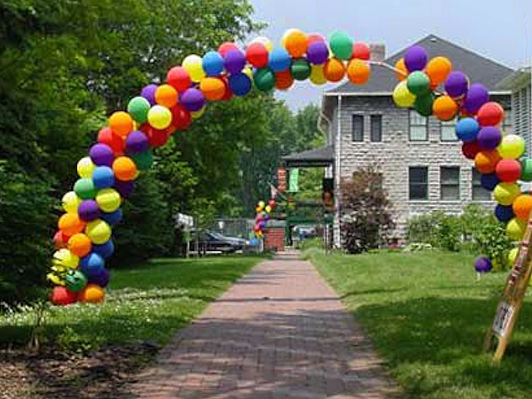 A colorful arch of balloons in various colors decorates a brick walkway leading to a house, with green grass and trees on either side. More balloons are visible near the house in the background.