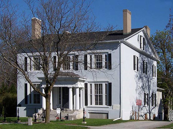 A large, two-story white house with black shutters, prominent chimneys, and a columned front porch stands behind leafless trees on a sunny day.