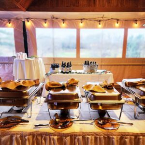 A buffet table set for an event, featuring covered chafing dishes with serving utensils in the foreground and a drink station with bottles and cups in the background, under warm string lights inside a tent.