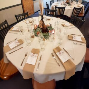 Round table set for a formal event with beige napkins, menus, glassware, and cutlery. A rustic centerpiece with greenery, candles, and a wooden table number 9 decorates the middle. Black chairs surround the table.