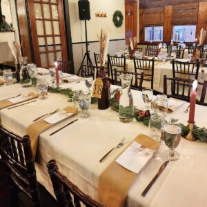 A banquet table set for a formal event with white tablecloths, tan runners, glassware, menus, candles, greenery, and numbered brown bottles. Wooden chairs are arranged around the table in a warmly lit room.