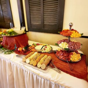 A buffet table with assorted crackers, cubed cheese, rolled deli meats, a bowl of dip, and fresh fruit, all arranged on wooden boards and tiered trays, covered with a white tablecloth.